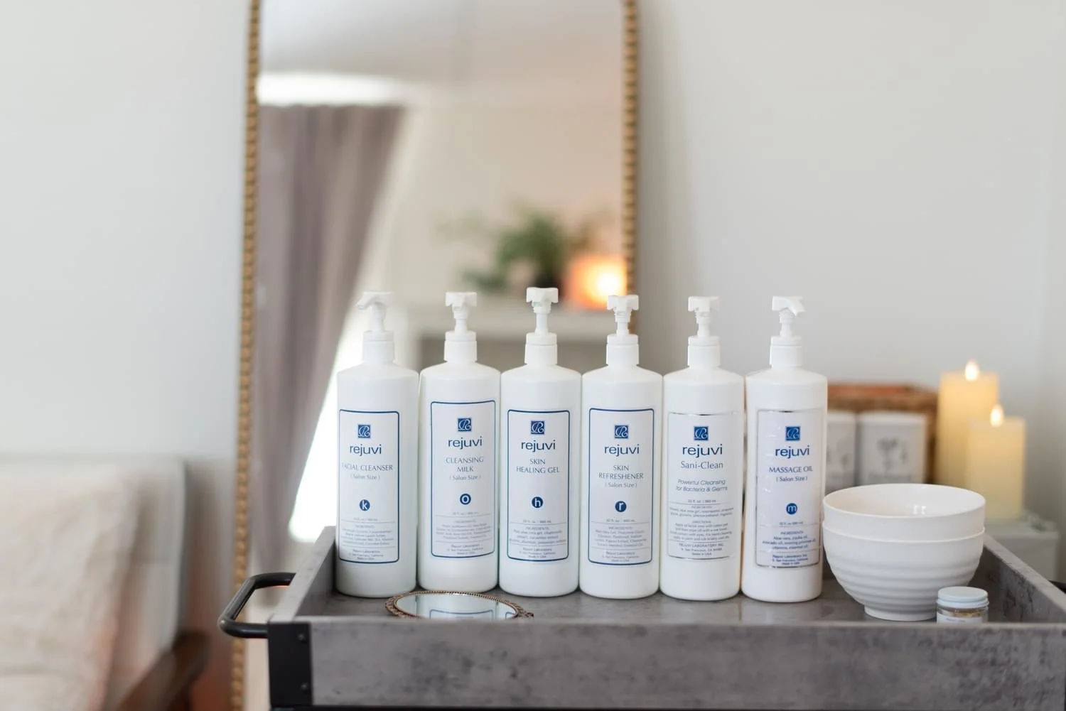 A row of Rejuvi pump bottles on a studio cart beside white bowls and lit candles