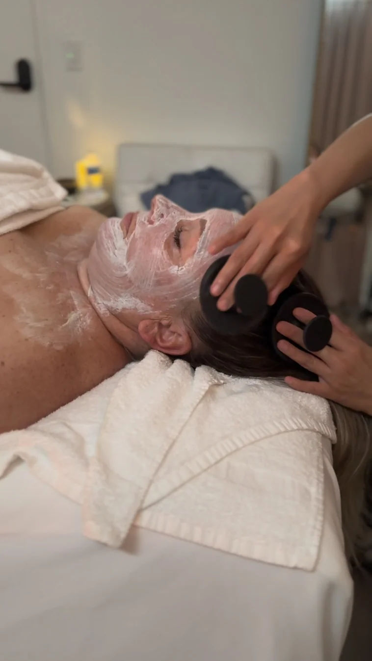 A client relaxes with a white mask on her face and chest while an esthetician's hands rest near her hairline