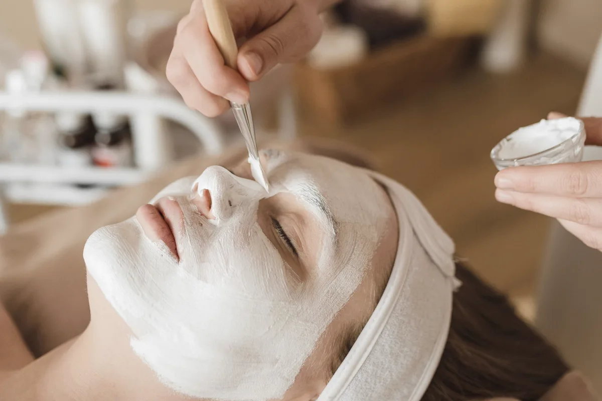 An esthetician brushes a white clay mask onto a client's face.