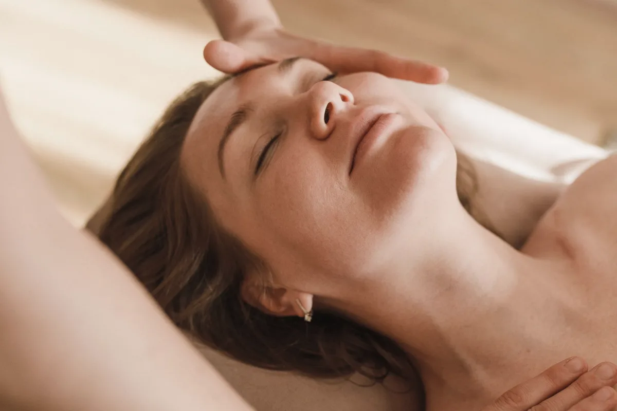 An esthetician's hands rest on a relaxed woman's forehead and collarbone during a facial massage.